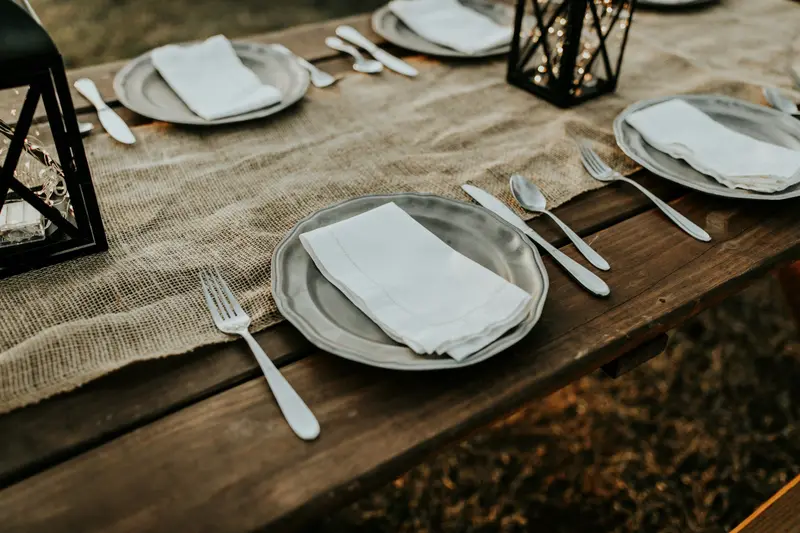 Une longue table de bois dressée : chemin de table en lin, assiettes, couverts et serviettes pliées.